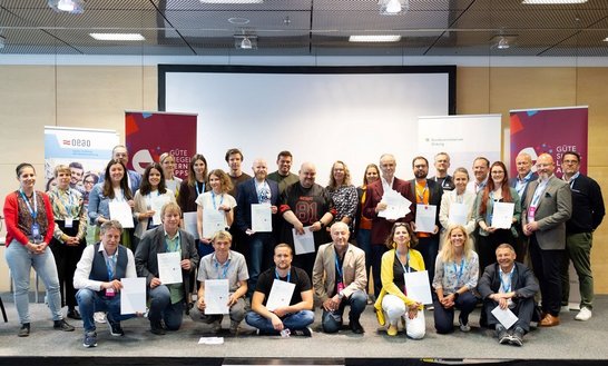 Gruppenbild mit ca. 35 Personen auf einer Bühne, die meisten halten ein Zertifikat in der Hand